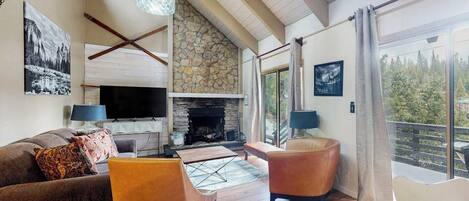 A living room with a stone fireplace, a television, and a large window overlooking a forest.