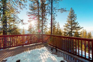 A wooden deck with a red railing overlooking a snowy landscape with tall pine trees.