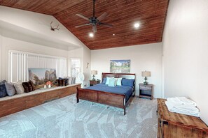 A bedroom with a wooden ceiling, a large bed, a bench with cushions, a window with blinds, and a painting on the wall.