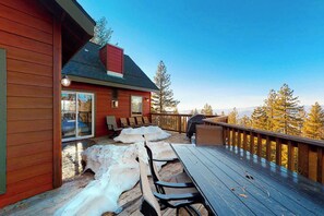 Una terraza de madera con mesa y sillas, una parrilla y vistas a árboles.