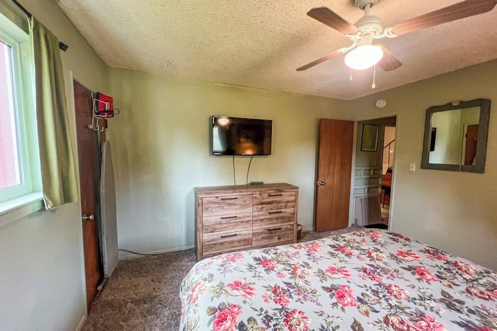 A bedroom with a bed, a ceiling fan, a dresser, a television, and a door leading to another room.