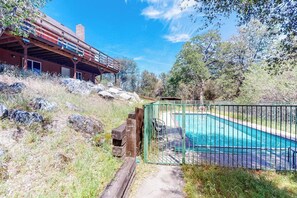 A pool area with a wooden deck, a fenced pool, and a building with a red roof in the background. - Hodges Hill Yosemite House (Oakhurst)