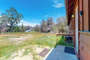 A building with a wooden exterior, a porch with a bench, and a view of a grassy area with trees. - Hodges Hill Yosemite House (Oakhurst)