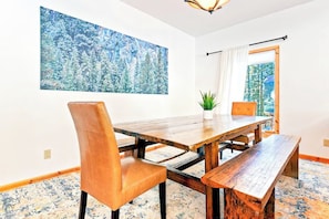 A dining area with a wooden table and chairs, a large painting on the wall, and a window with curtains. - Oso Lodge - Ping-Pong, Deck & EV Charger (Yosemite West)