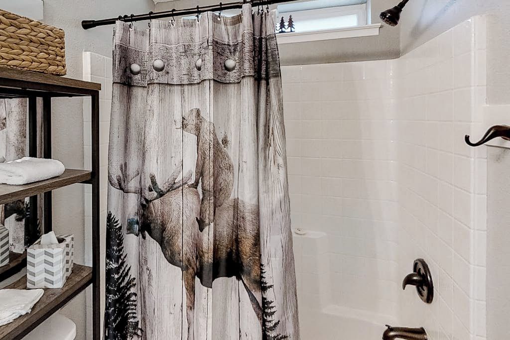 Un baño con cortina de ducha con diseño de alce, estante de madera con artículos de aseo y bañera con grifo.