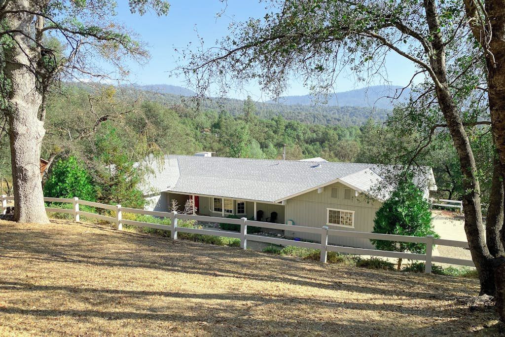 Una casa a un piano con tetto spiovente, circondata da un recinto bianco, sullo sfondo di alberi e colline.