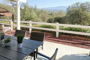 Eine Terrasse im Freien mit Tisch und Stühlen, umgeben von einem weißen Zaun, mit Blick auf einen angelegten Garten und sanfte Hügel in der Ferne.