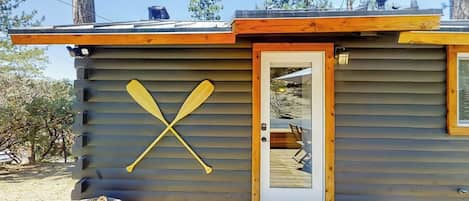 Una cabaña con puerta de madera, una terraza y una pala apoyada en la pared.