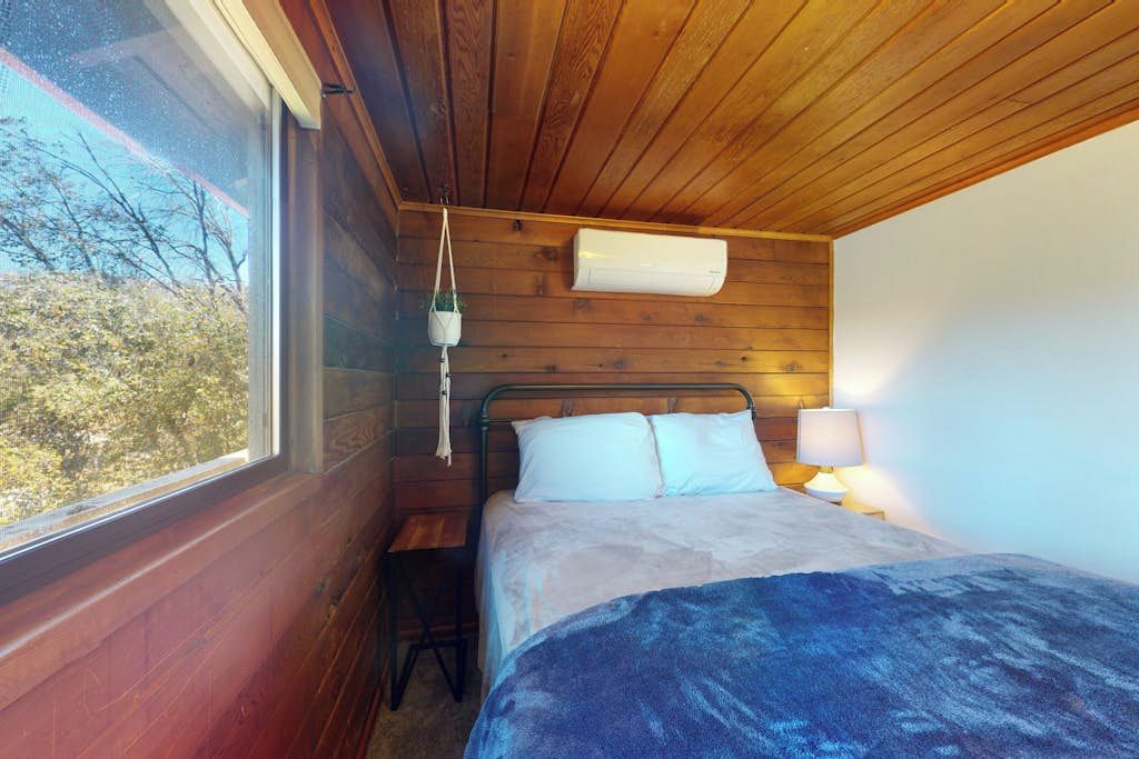Quarto com cama, mesa de cabeceira, luminária, planta pendente e janela.