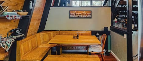 A cozy wooden interior with a bench seating area, a sign reading 'Welcome to our NECK OF THE WOODS,' and a window providing a view of trees.
