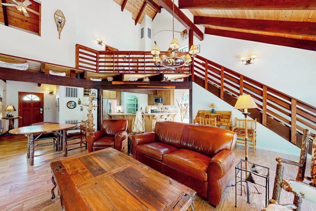 Ampla sala de estar com escada de madeira, mobiliário de couro e cozinha ao fundo.