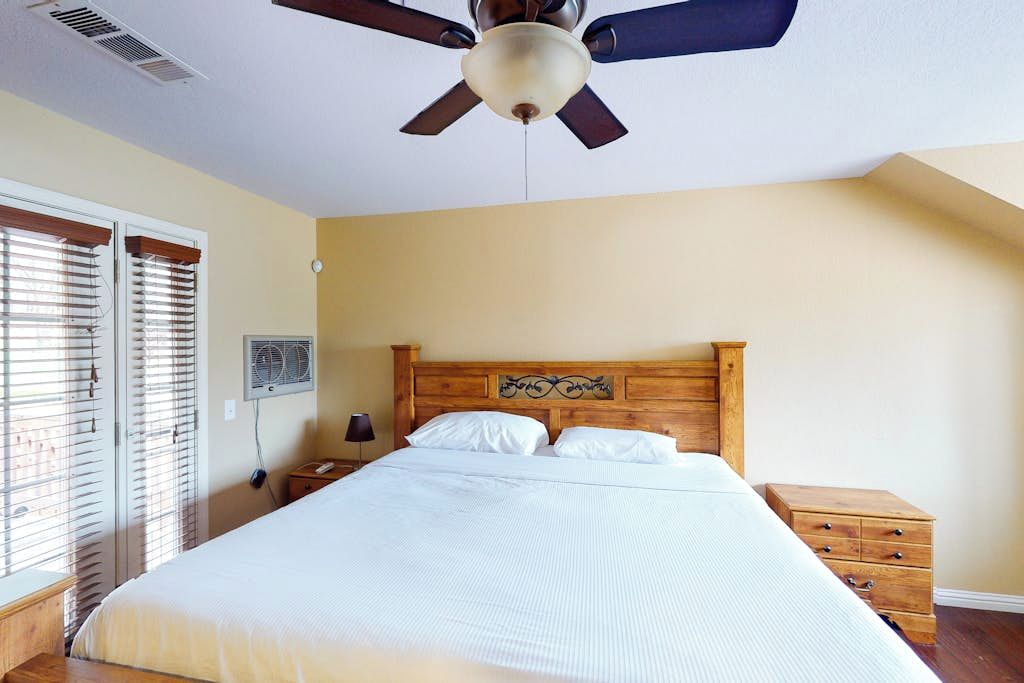A bedroom with a bed, wooden headboard, bedside tables, a ceiling fan, and a window with blinds.