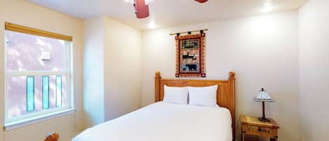Quarto com cama, ventoinha no teto, mesa de cabeceira com abajur, janela com persianas e um objeto decorativo na parede.