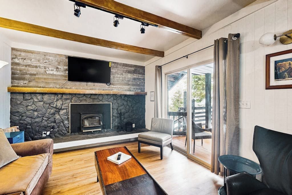 Uma sala de estar moderna com lareira de pedra, vigas de madeira, televisão de tela plana e uma grande porta de vidro deslizante que dá para uma área externa.
