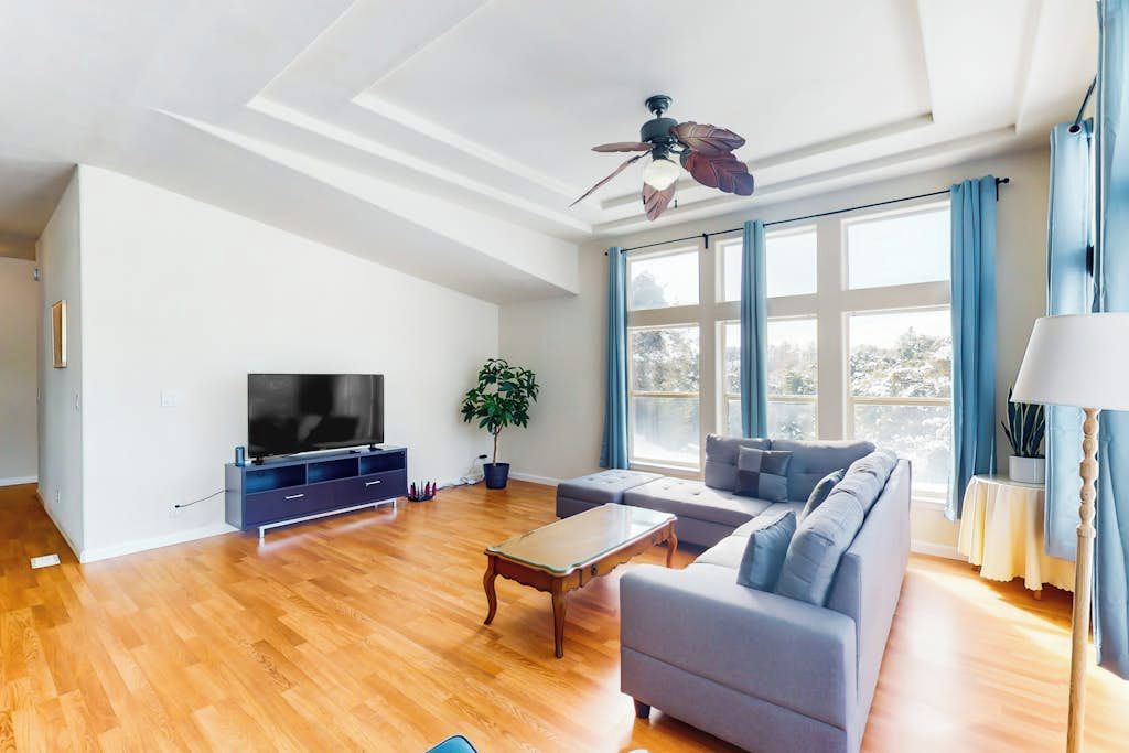 A spacious living room with a flat-screen TV, a ceiling fan, a large window with curtains, and a sofa.