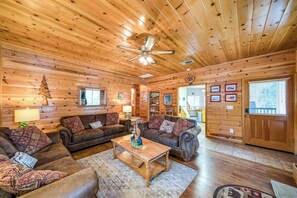 A log cabin interior with a central seating area, a ceiling fan, and a door leading to another room. - Black Bear Cabin - Boat Slip, Walk to Lake (Bass Lake)