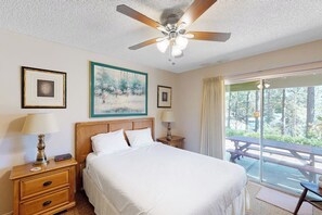 A hotel room with a bed, two bedside tables, a ceiling fan, and a large window with a view of a forest. - Miglin Lakeview - Lake Views & Private Boat Slip (Bass Lake)