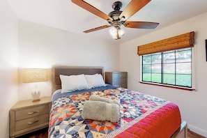 Quarto com cama, mesa de cabeceira, luminária, ventoinha no teto, janela com persianas e vista para o exterior.