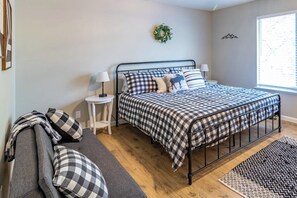 A bedroom with a metal bed frame, checkered bedding, a small bedside table with lamps, a grey sofa, and a window with a view. - Pine Place - Near Bass Lake & Yosemite (Oakhurst)