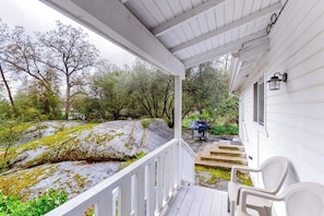 A covered porch with a white railing, a stone pathway leading to a grill area, and a view of a natural landscape with trees and rocks. - Pine Place - Near Bass Lake & Yosemite (Oakhurst)