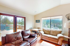A living room with a brown leather sofa, a beige sofa, a dining table with chairs, and a large window with a view of trees. - Alder Lake View Lodge (Bass Lake)
