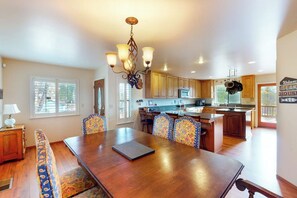 A spacious dining area with a wooden table and patterned chairs, a kitchen with wooden cabinets, and a hanging light fixture. - Alder Lake View Lodge (Bass Lake)