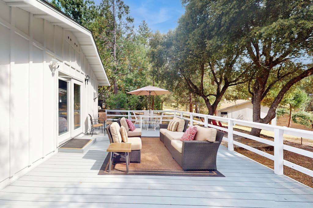 A covered outdoor deck with wicker furniture and a table, surrounded by a white fence and trees.