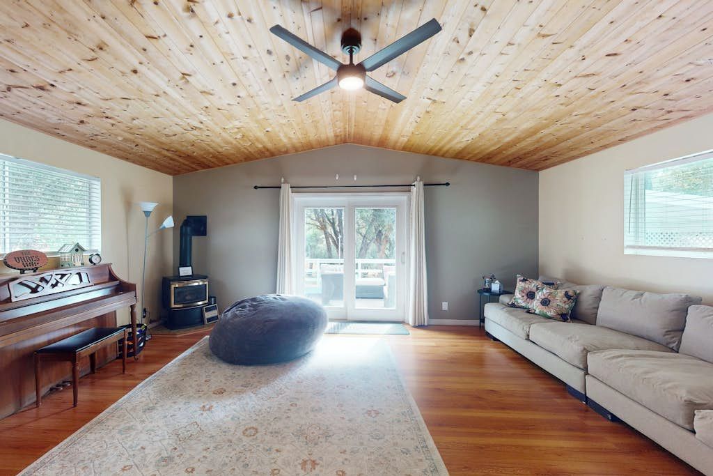 A spacious room with a wooden ceiling, a piano, a bean bag chair, a sofa, and a large window.