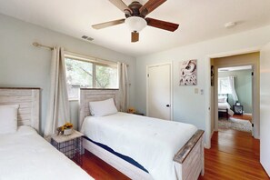 Une chambre avec deux lits, un ventilateur de plafond, une fenêtre avec des rideaux et une porte donnant sur une autre pièce.