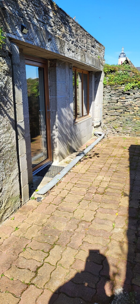 Edifici de pedra amb una finestra i una porta, zona pavimentada amb gespa, i una construcció a la distància.