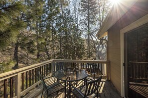 Eine Holzterrasse mit einem Glastisch und Stühlen, umgeben von Bäumen, und eine Tür rechts.
