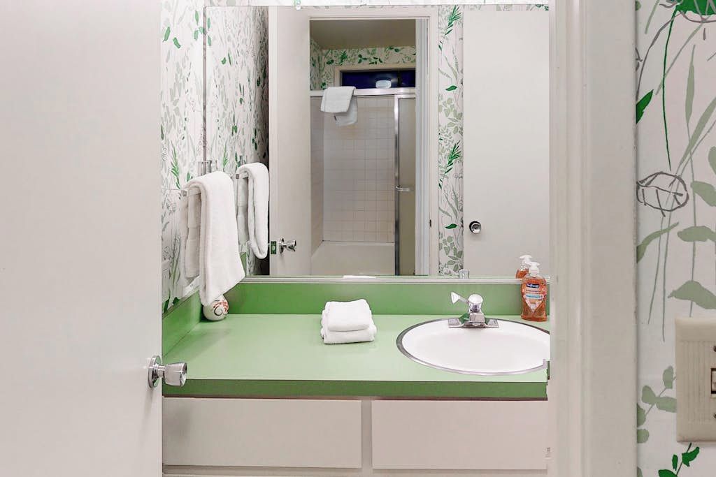 Uma casa de banho com bancada verde, lavatório branco, espelho e chuveiro visível através de uma porta aberta.