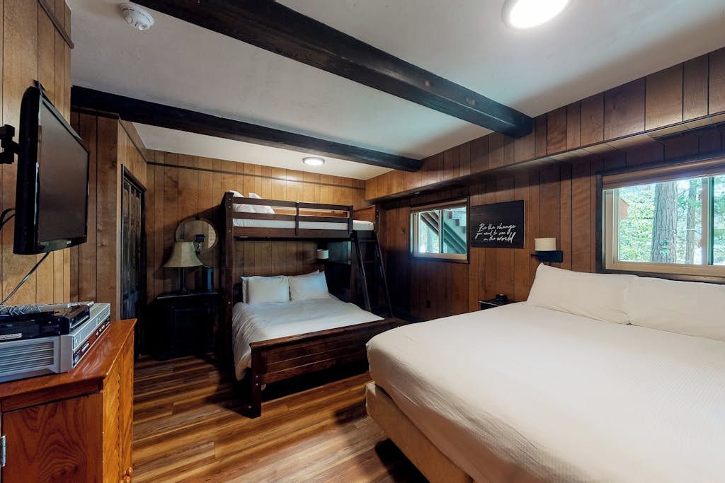 A room with wooden walls, a bunk bed, a flat bed, a desk with a computer, and a window with a view of trees.