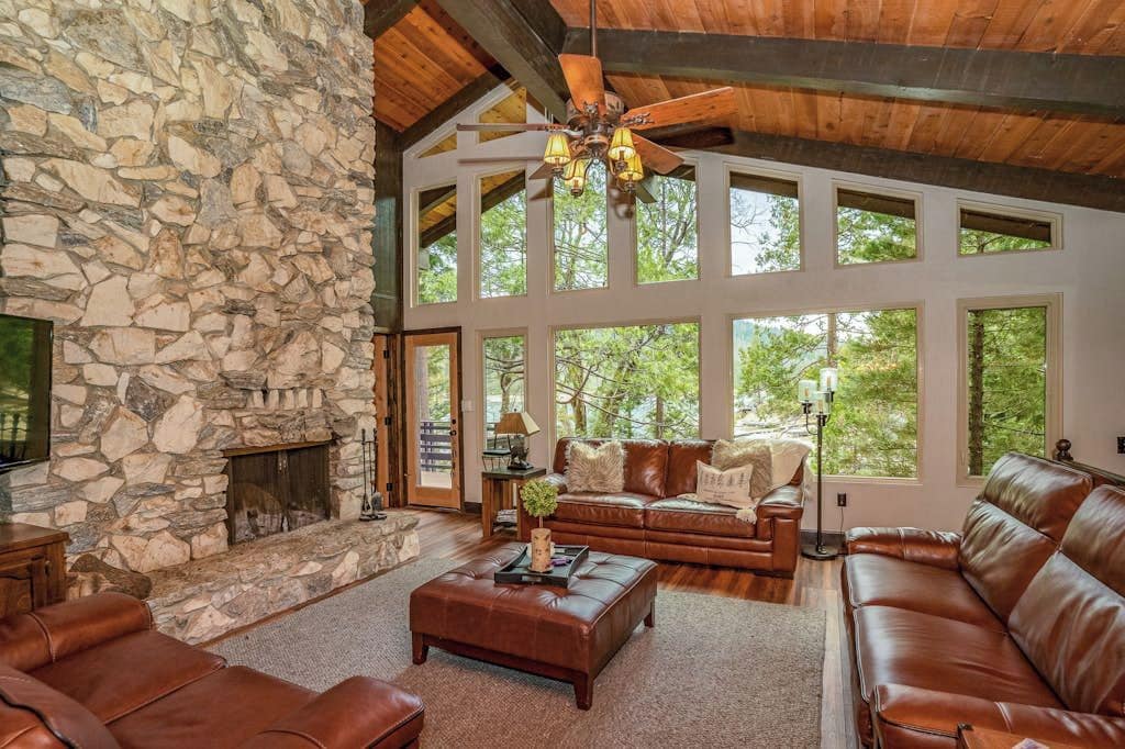 A spacious living room with a stone fireplace, leather sofas, and large windows offering a view of the outdoors.