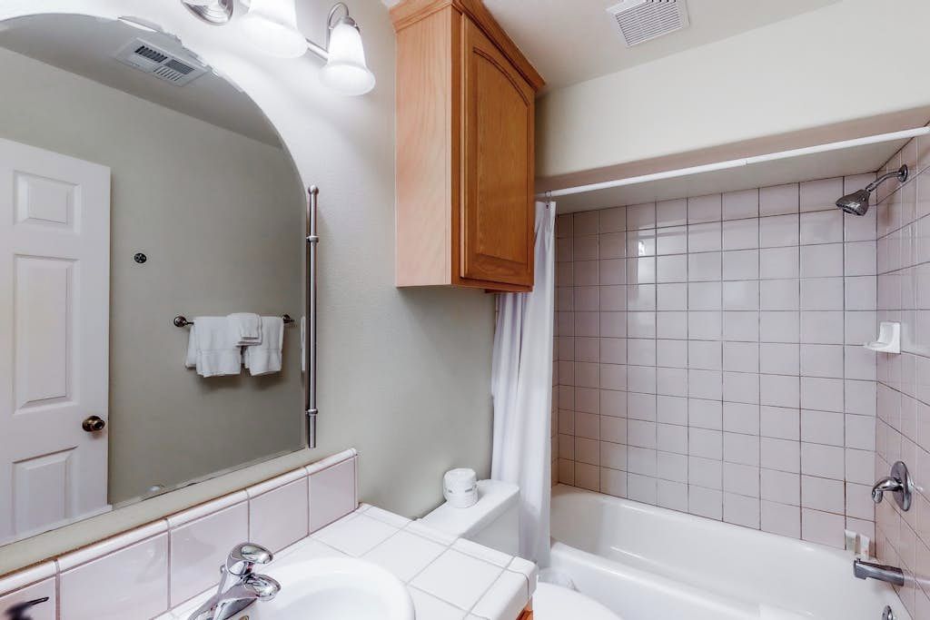 Ein Badezimmer mit weißem Waschbecken, Spiegel und Badewanne. Oberhalb des Spiegels befindet sich ein Holzkästen und über der Badewanne ein Duschvorhang.
