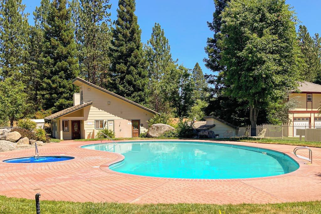 Una piscina con solárium de ladrillo, rodeada de árboles y un edificio pequeño.