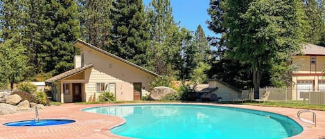 Una piscina con solárium de ladrillo, rodeada de árboles y un edificio pequeño.