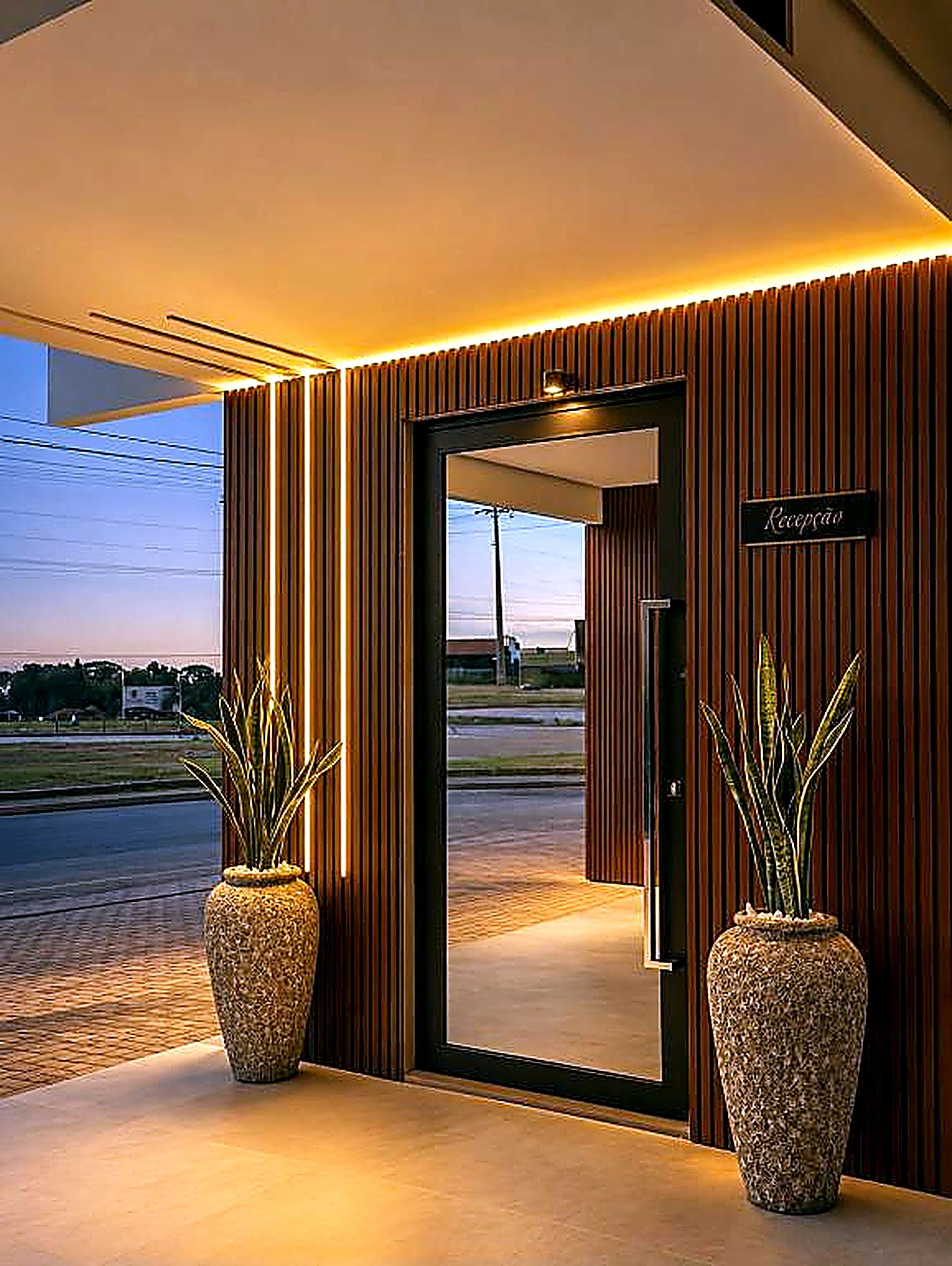 Entrada de un hotel con una recepción, dos plantas grandes en macetas y vista a la calle.