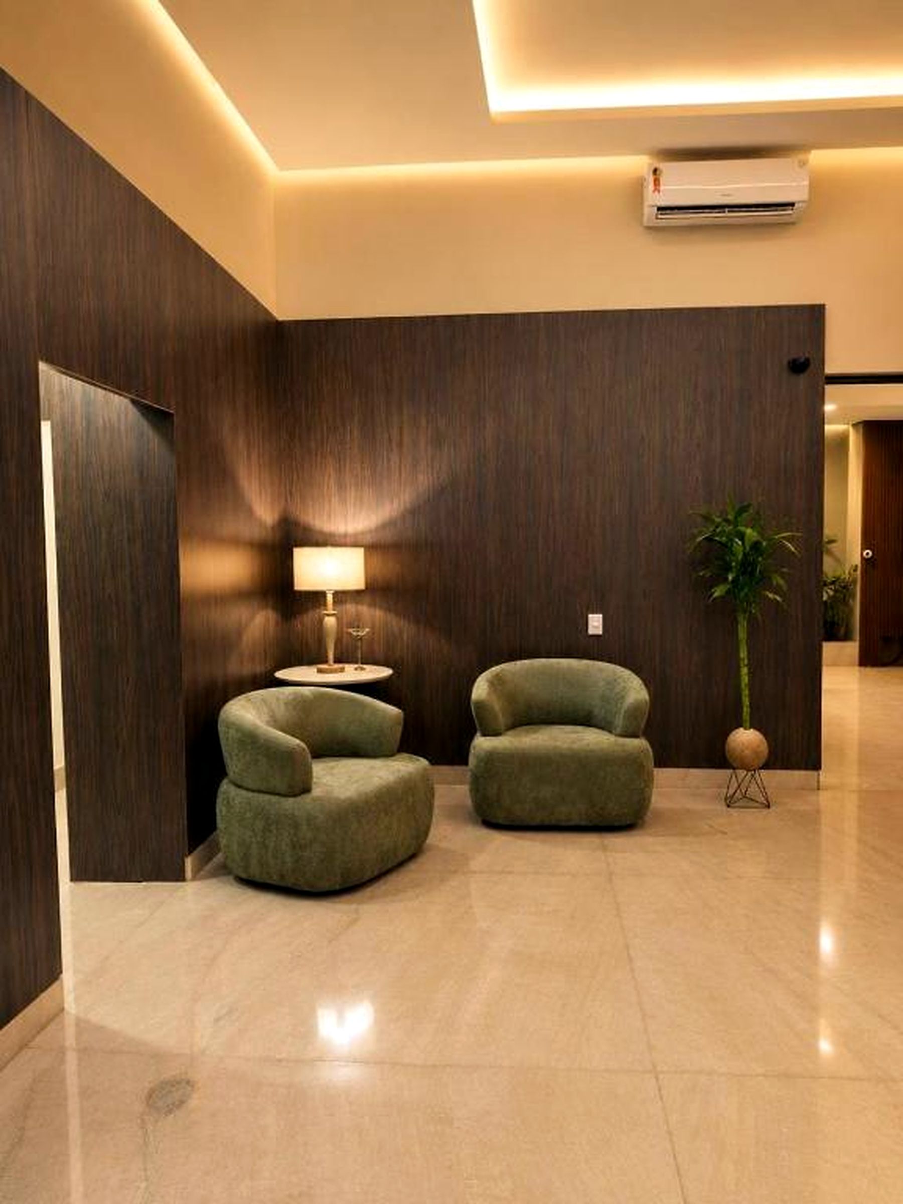 A modern lobby with two green armchairs, a floor lamp, a potted plant, and an air conditioning unit.