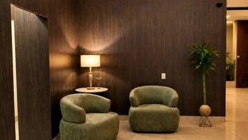 A modern lobby with two green armchairs, a floor lamp, a potted plant, and an air conditioning unit.