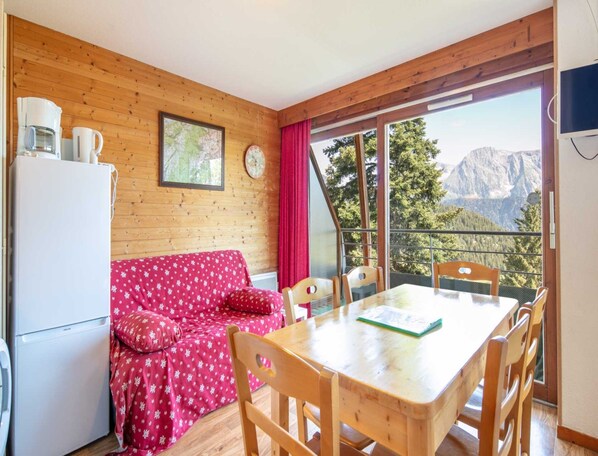A cozy room with a wooden interior, a red sofa, a dining table with chairs, a refrigerator, and a large window offering a mountain view. - Alpine Ski Apartment in Tignes for 4 (Chamrousse)