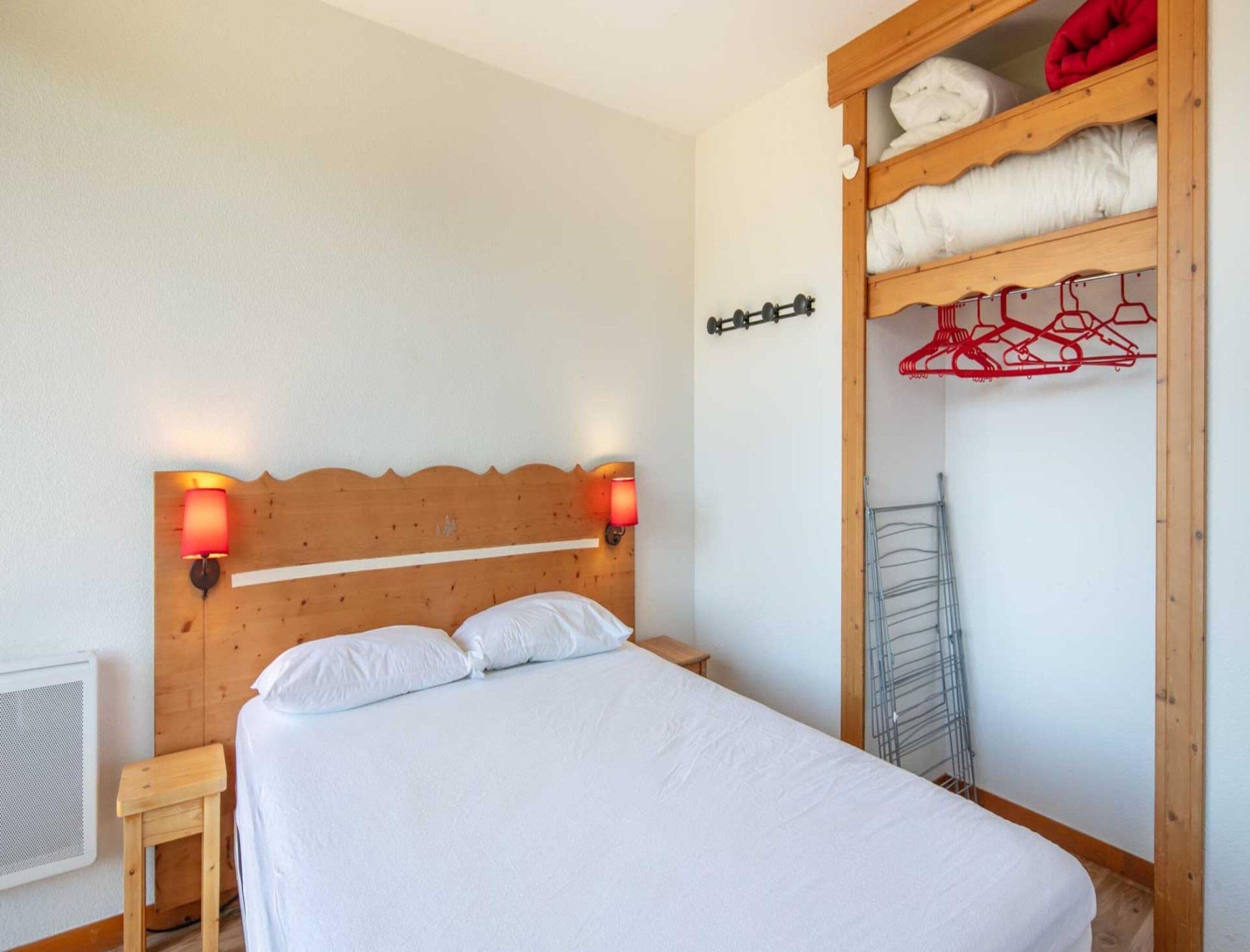 Un lit simple avec une tête de lit en bois, deux lampes fixées au mur, une petite table de chevet et une armoire intégrée avec un étendoir à linge.