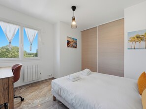 A bedroom with a bed, a desk, and a chair. There is a window with curtains, a radiator, and a picture on the wall. - Le Solévin · Vue vignes & Pic Saint-Loup - 4p (Jacou)