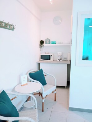 Ett litet, praktiskt kök med mikrovågsugn, kaffebryggare och en liten hylla. Där finns en vit stol med en turkosfärgad kudde och ett litet bord med ett kort på.