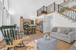A modern living room with a grey sofa, a rocking chair, a dining table with chairs, and a staircase with a wooden railing.
