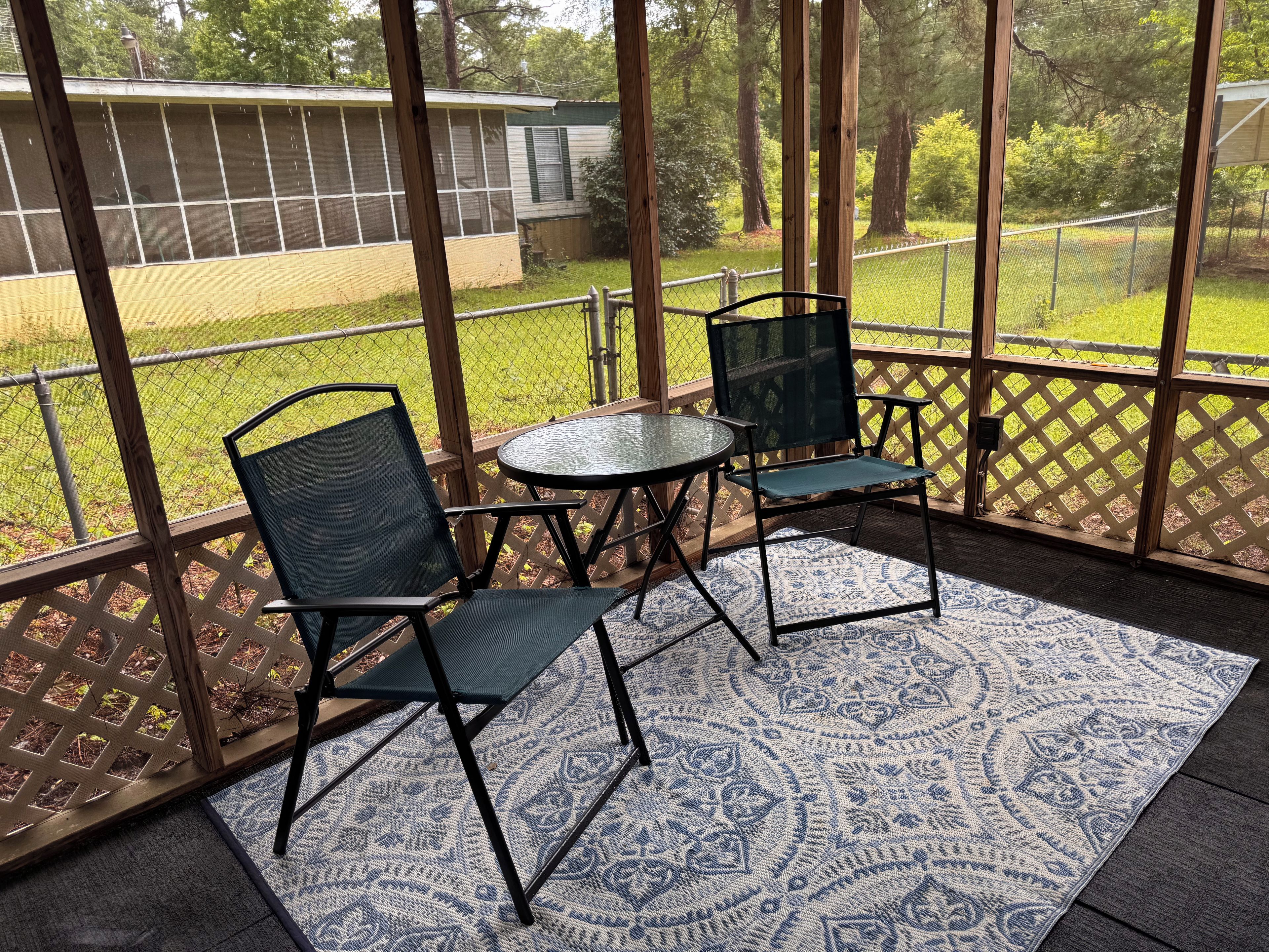 A covered patio with two chairs and a table, a patterned rug, and a view of a house through a wooden lattice.