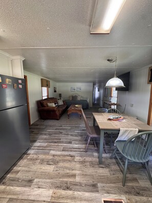 A compact kitchen with a refrigerator, a dining table with chairs, a wooden floor, and a living area with a sofa and a coffee table.