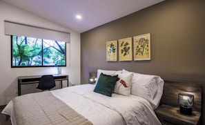Ein Schlafzimmer mit Bett, Nachttisch, Lampe, Schreibtisch, Stuhl und Fenster mit Blick auf Bäume.