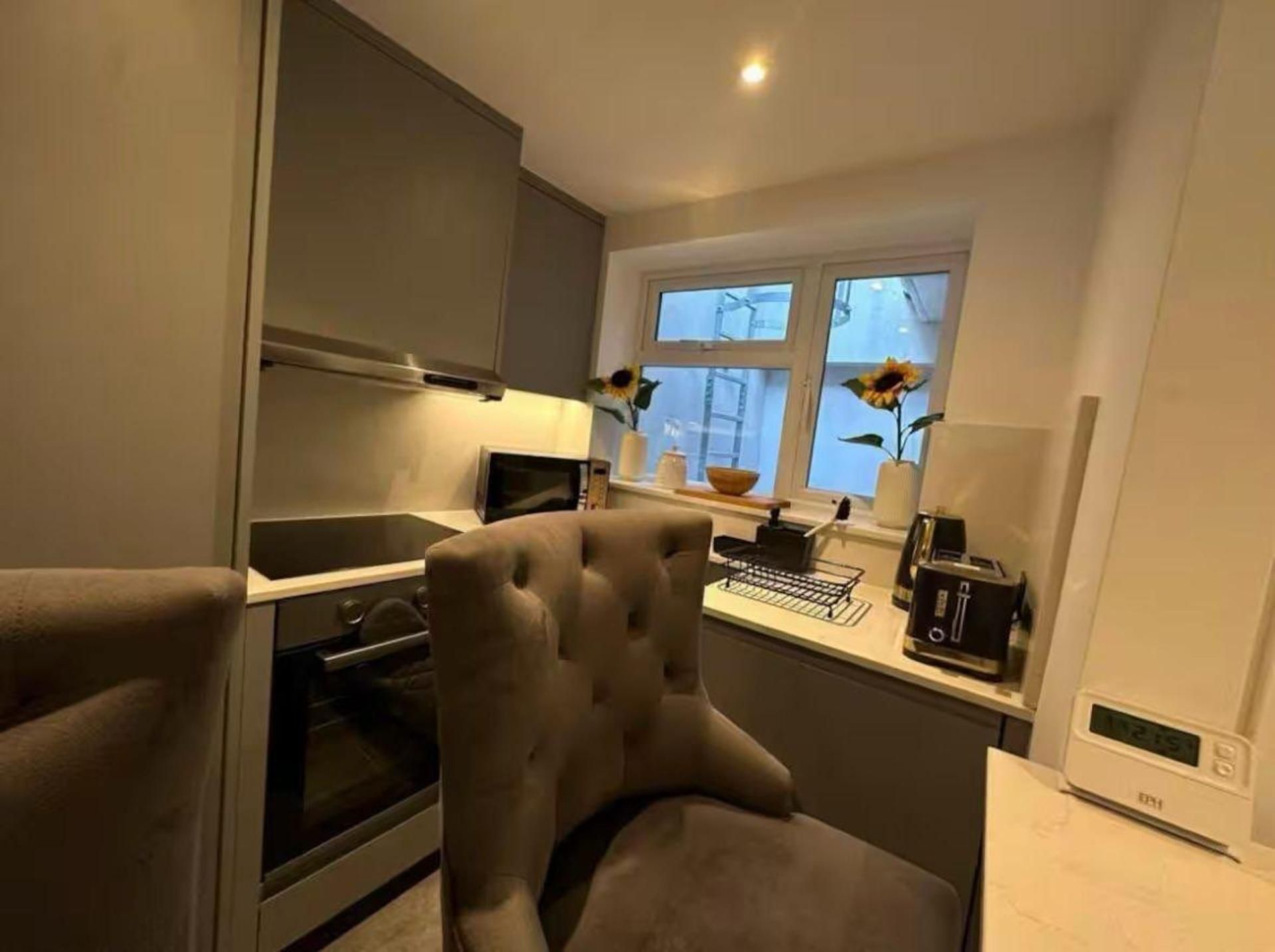 Cocina compacta con horno integrado, microondas y tostadora sobre la encimera. Hay una ventana con vistas al exterior y una silla frente a la encimera.