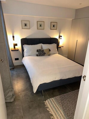 En pænt redt seng med hvide sengetøj og dekorative puder, et sengebord med lampe, og en gulvtæppe med et mønster på gulvet.
